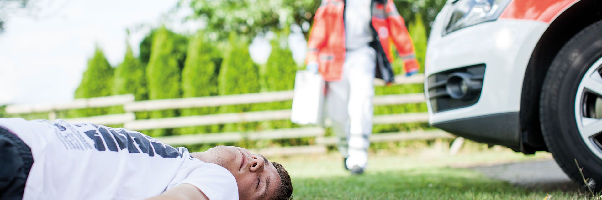 Foto: Ein junger Mann liegt bewusstlos auf einer Wiese. Ein DRK-Mitarbeiter mit Koffer nähert sich dem Patienten. Am Rande des Fotos ist ein Fahrzeug des Deutschen Roten Kreuzes zu sehen.