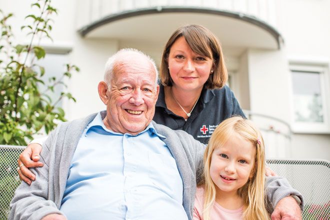 Foto: Ein Mädchen, ein älterer Herr und eine DRK-Mitarbeiterin blicken freundlich und warmherzig in die Kamera. Sie wirken zusammen wie ein Team.